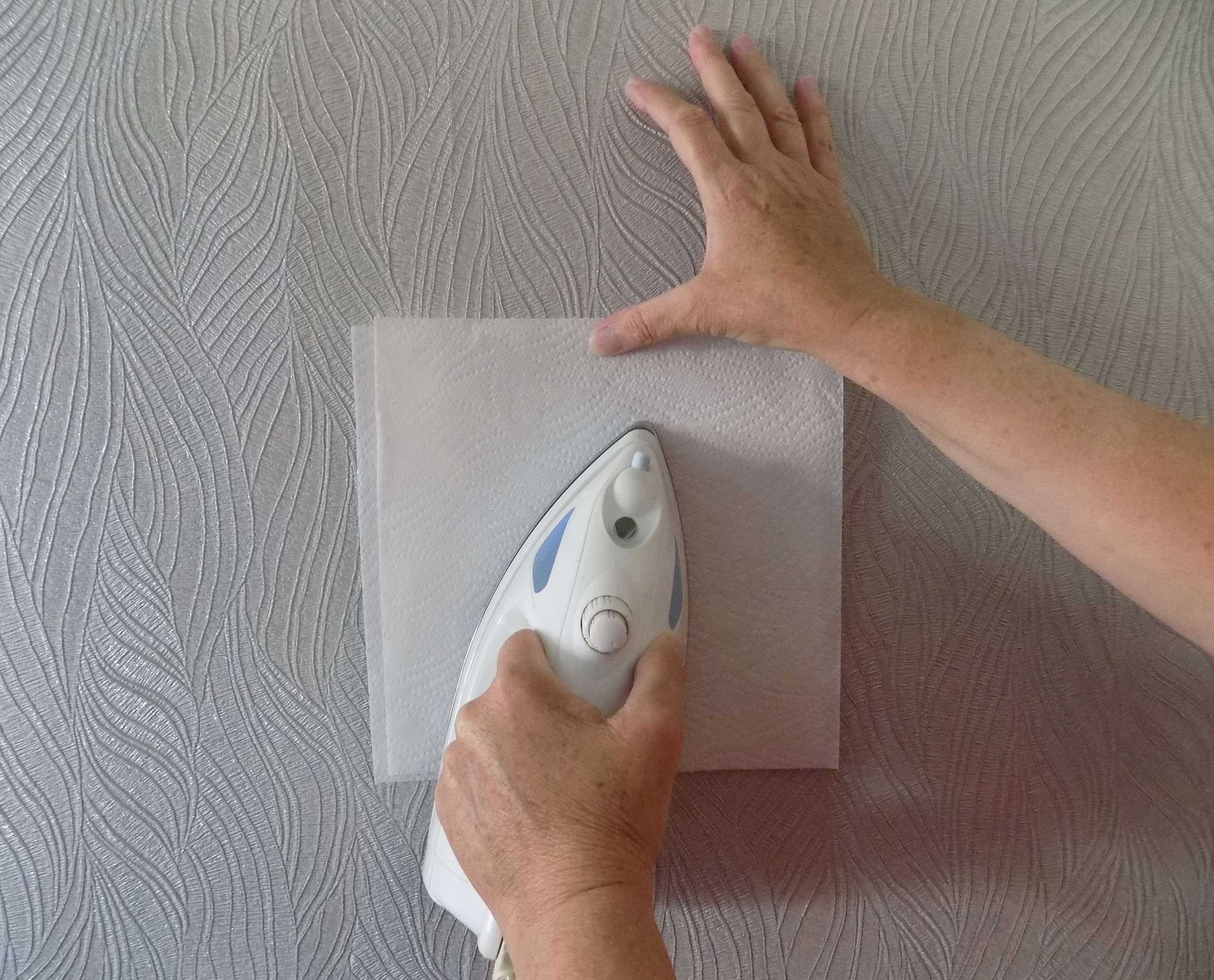 person holding iron and kitchen roll against wallpaper to clean off grease