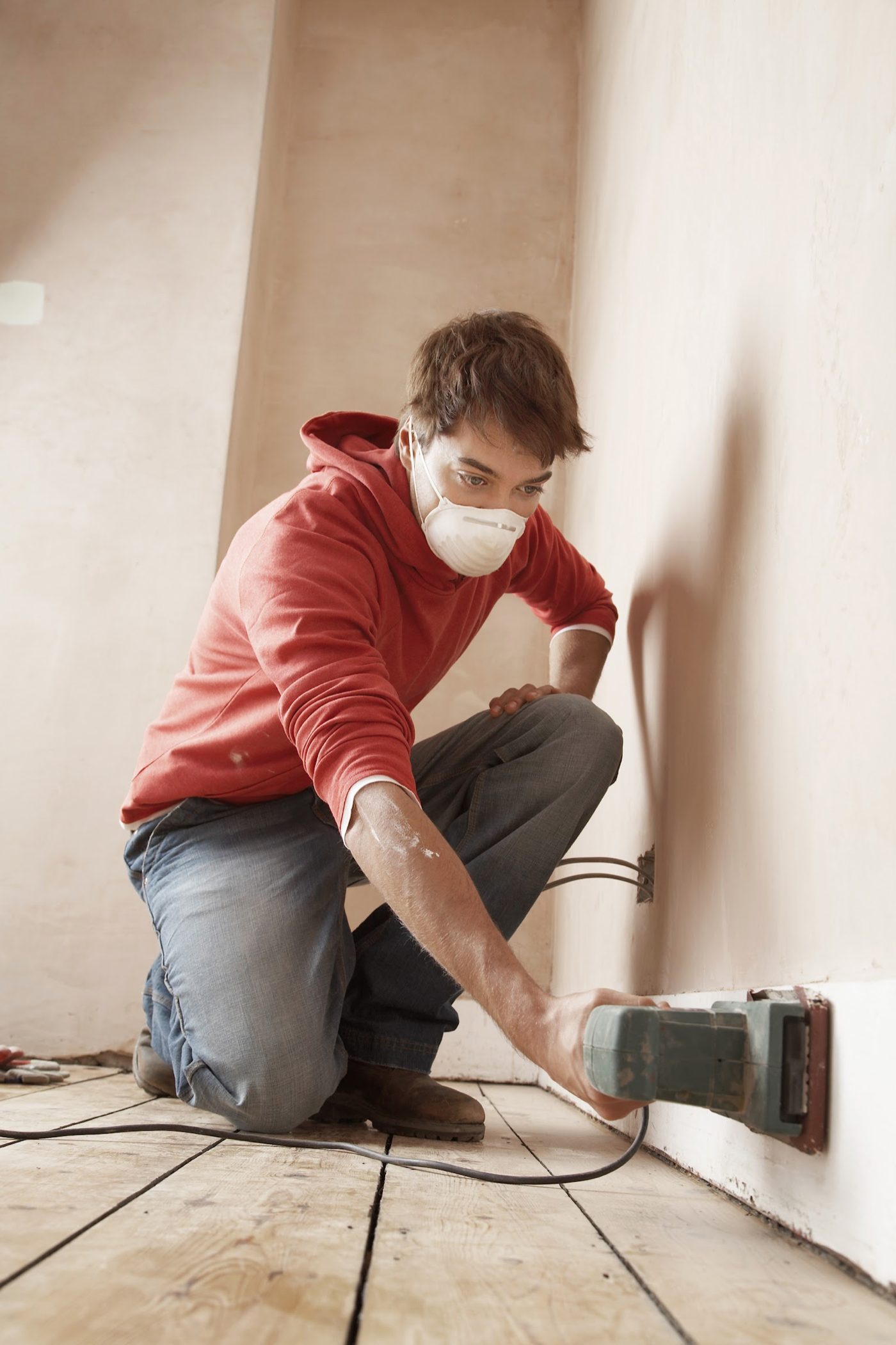 man sanding skirting board with electric sander in red hoody, jeans and face mask